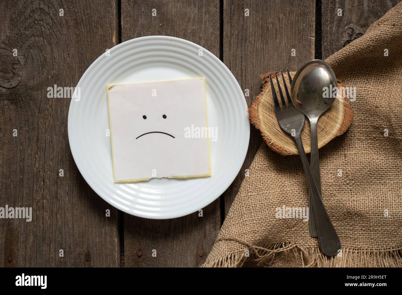 un sorriso triste si trova su un foglio di carta su un piatto accanto a un cucchiaio e una forchetta su uno sfondo marrone burlap, niente da mangiare, crisi Foto Stock