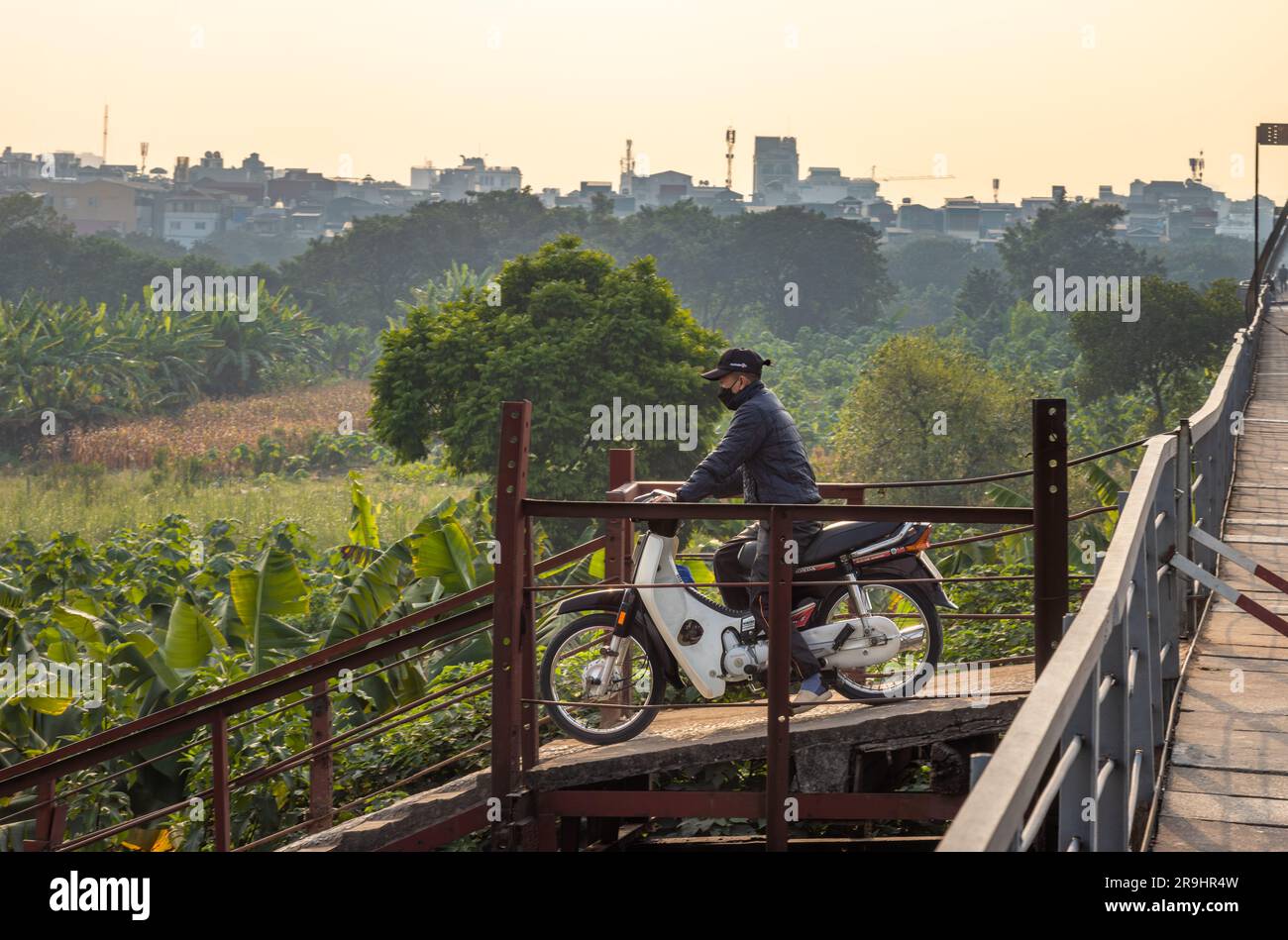 Un motociclista su uno scooter e indossa una maschera facciale e senza casco guida il Long Bien Bridge nello smog verso un'isola nel Red River, Hanoi, Vietna Foto Stock
