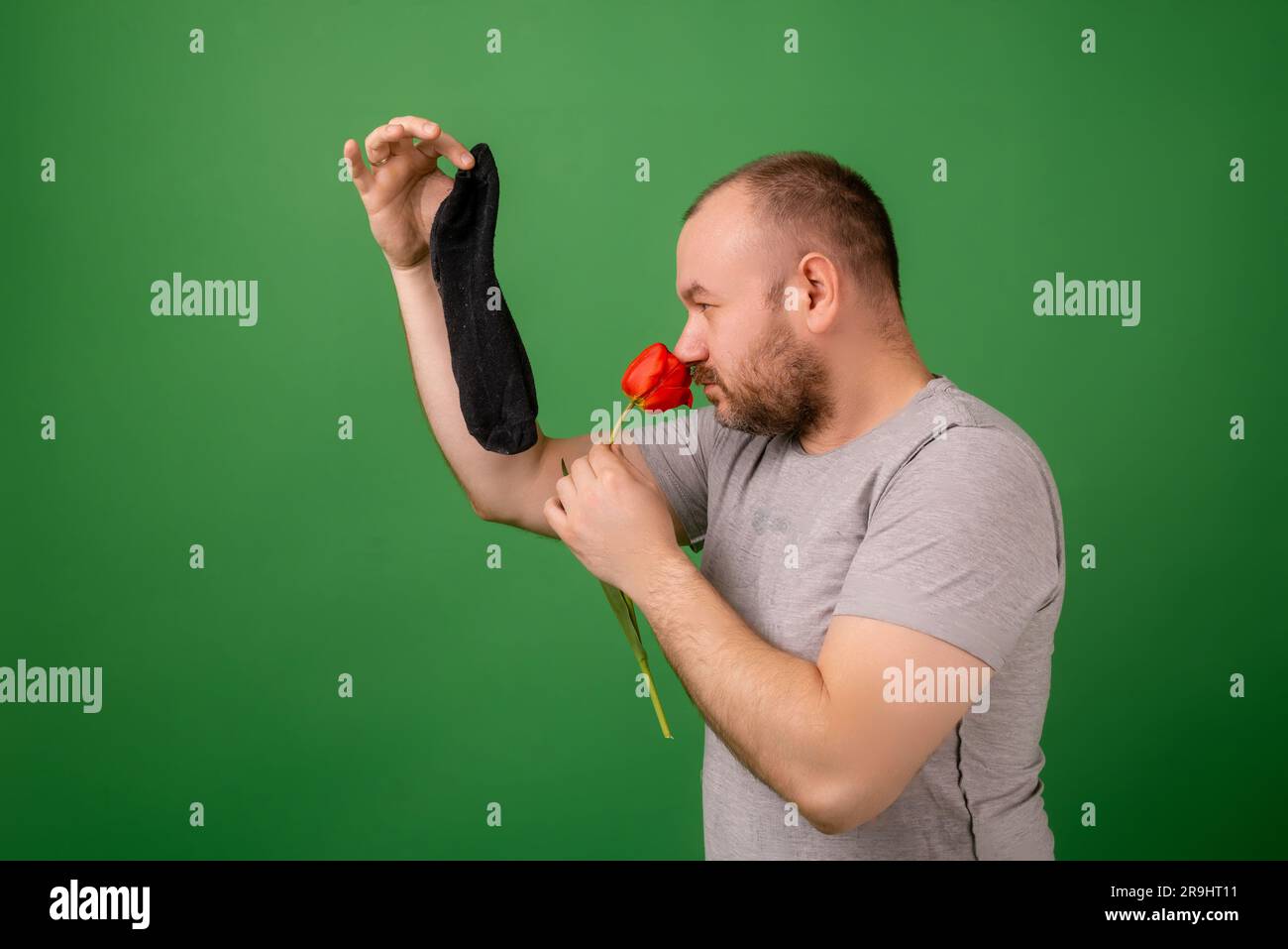 Un uomo di mezza età che regge un calzino sporco e puzzolente e un fiore fresco su sfondo verde. Lavanderia, igiene, odore di piedi. Foto Stock
