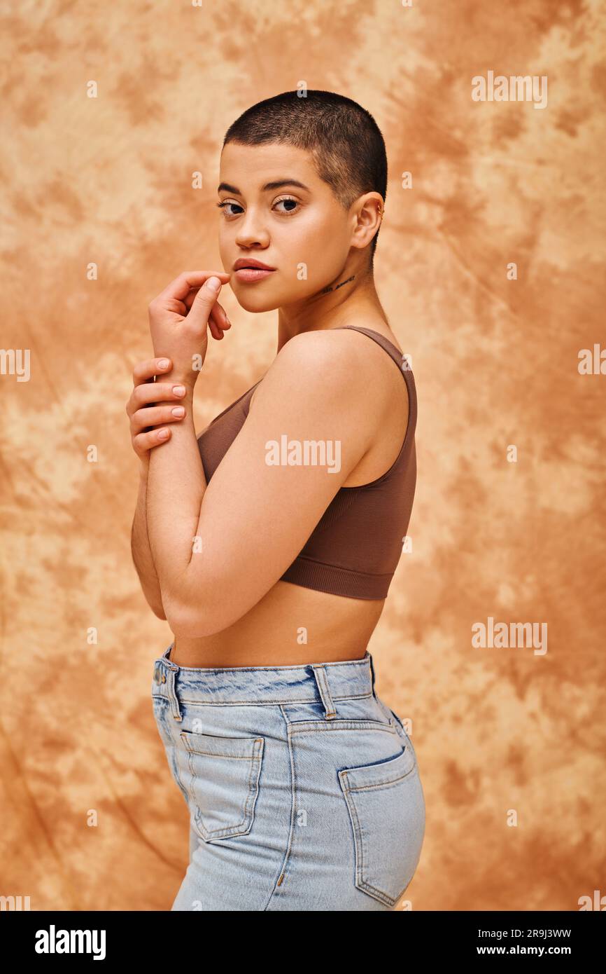 rappresentazione di corpo, curvy e giovane donna in top e jeans in posa su sfondo beige macchiato, pelo corto, auto-accettazione, generazione z, t Foto Stock