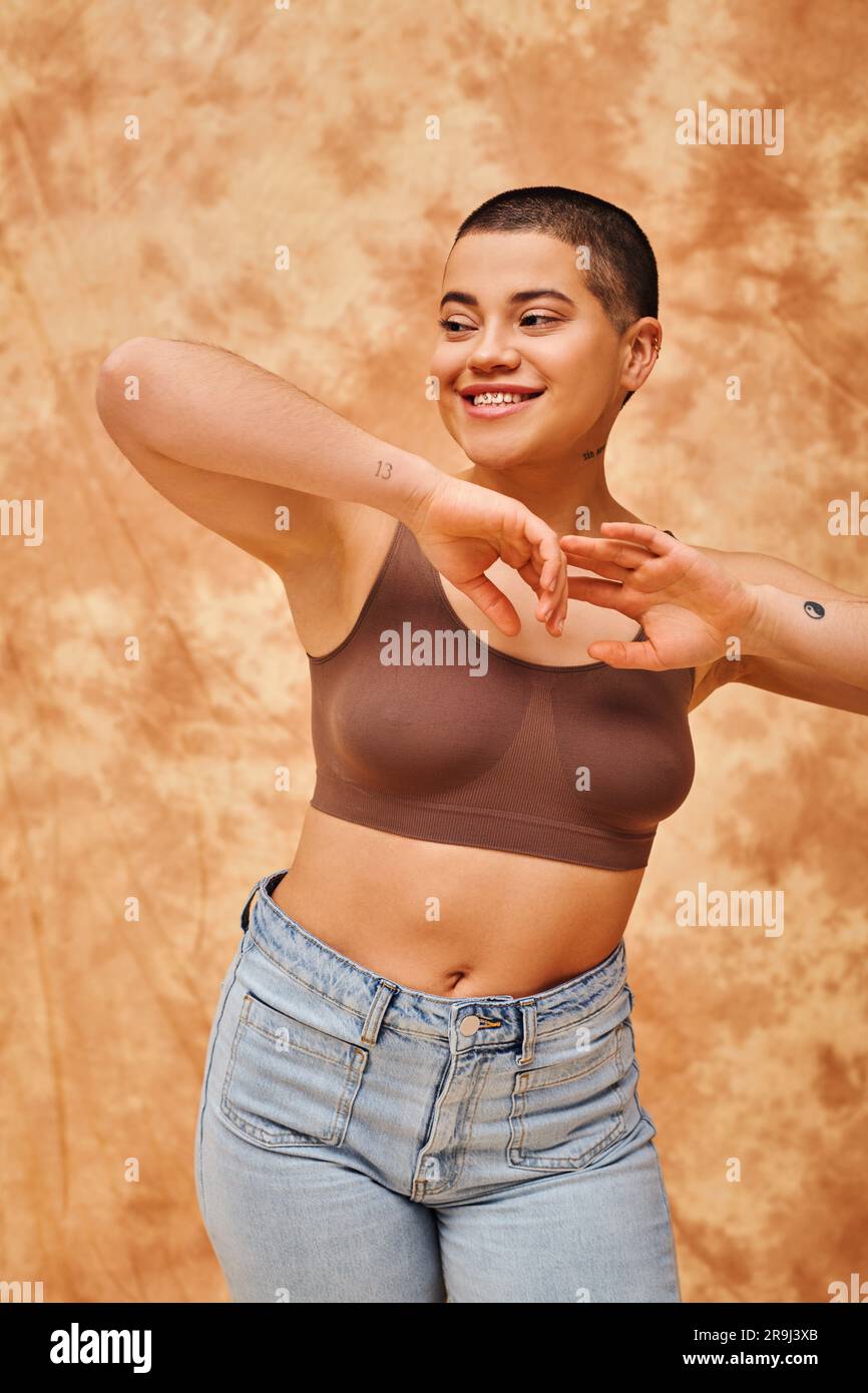 positività corporea, donna gioiosa e curvata in top e jeans in posa su sfondo beige chiazzato, posa yoga, auto-accettazione, generazione z, tatuata, Foto Stock