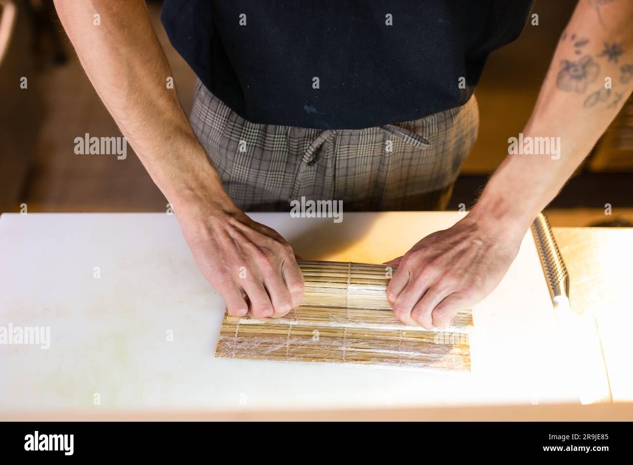 Dall'alto di uno chef maschile tatuato inriconoscibile in grembiule vicino al tavolo e che prepara sushi roll con tappetino di bambù con le mani mentre lavora in cucina Foto Stock