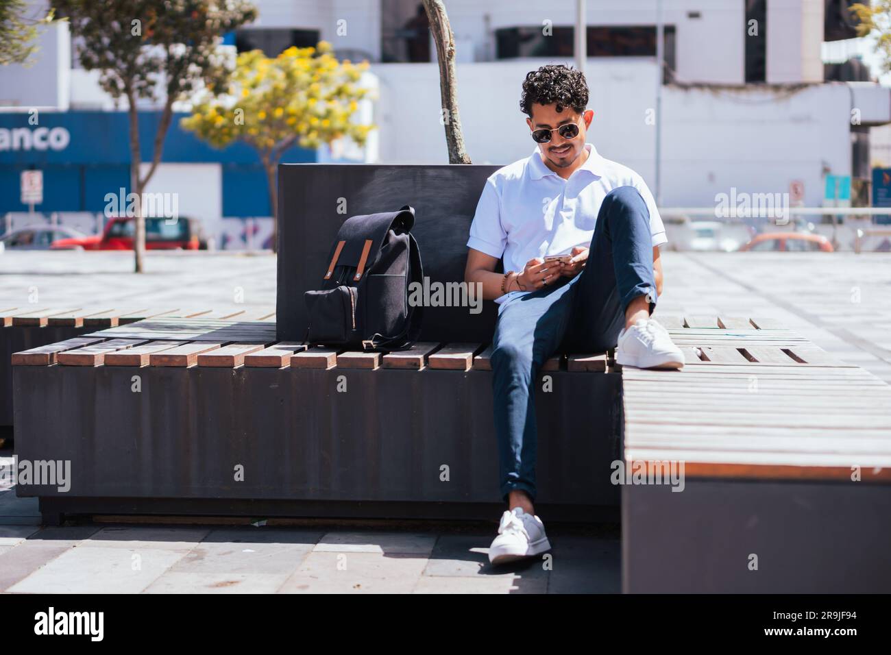 Corpo completo di uomo ispanico con occhiali da sole seduti sulla panchina e sfogliando il cellulare mentre riposi nelle giornate di sole in città Foto Stock