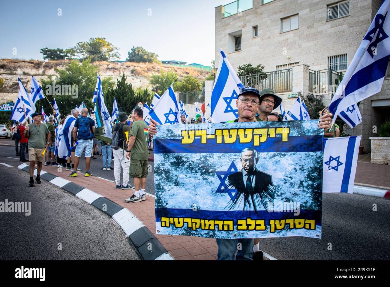 Modiin, Israele. 27 giugno 2023. Avinoam Poran 67, detiene un cartello che dice in ebraico: "Cattivo dittatore il cancro nel cuore della democrazia durante una protesta fuori dalla parte della casa del ministro della giustizia Yariv Levin. Centinaia di manifestanti contro la revisione giudiziaria del governo organizzarono una manifestazione di fuoco fuori dalla casa del ministro della giustizia Yariv Levin, mentre giurarono di intensificare le proteste sull'avanzamento di un disegno di legge per limitare il potere dell'alta Corte di giustizia di rivedere le decisioni del governo. (Foto di Eyal Warshavsky/SOPA Images/Sipa USA) credito: SIPA USA/Alamy Live News Foto Stock