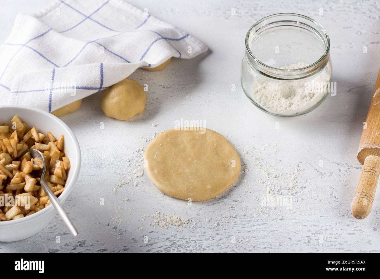 Pezzo piatto rotondo di impasto arrotolato, ripieno di mele con cannella, farina e mattarello su un tavolo grigio chiaro. Cucinare deliziose mini torte con la mela Foto Stock