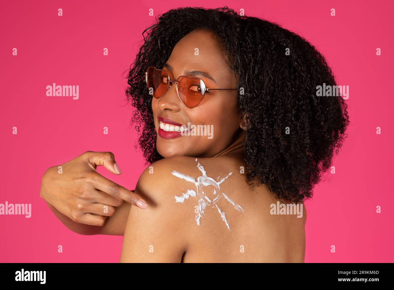 Felice giovane donna riccia nera con occhiali da sole che punta il dito contro la crema solare alle spalle Foto Stock