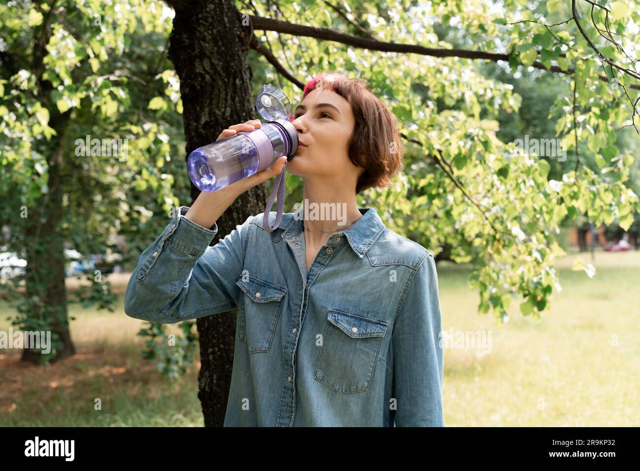 Una donna con i capelli rosa beve acqua nel parco dal caldo Foto Stock