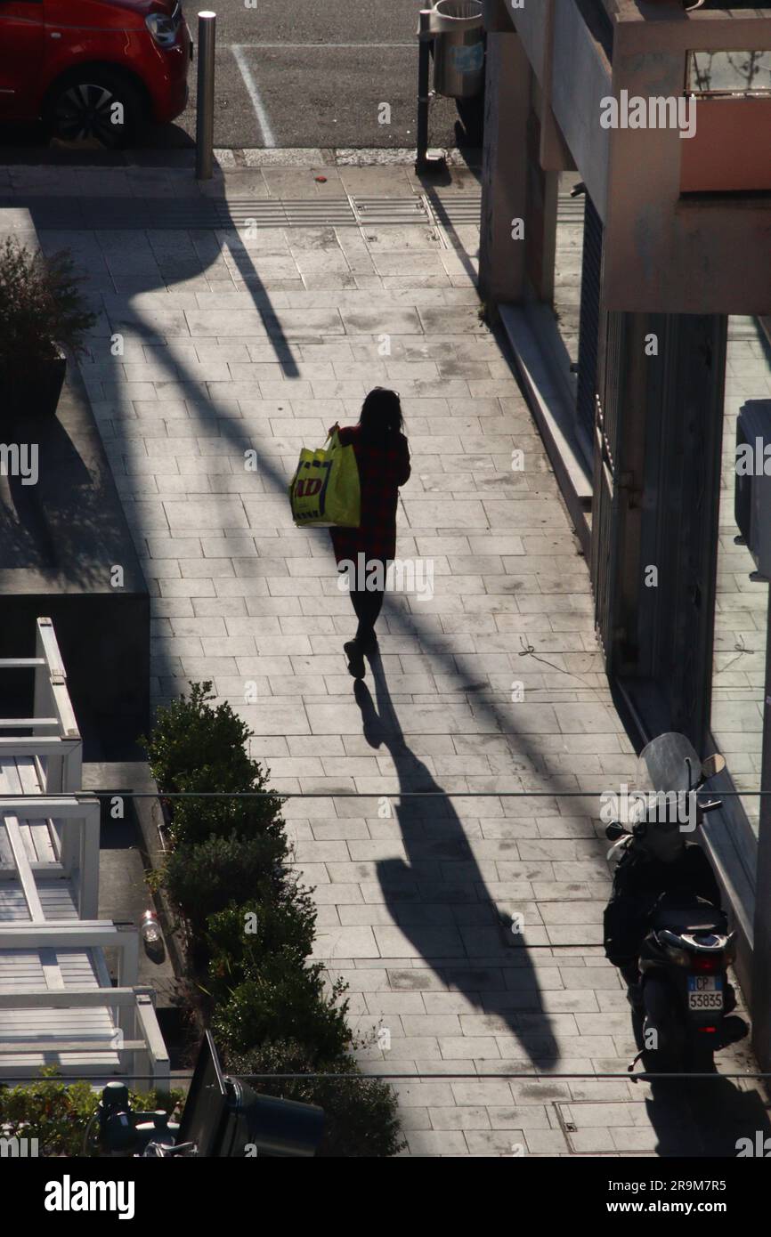 Una giovane donna lascia il suo posto di lavoro a casa, camminando al sole di sera, ignara della lunga ombra gettata dietro di lei, Messina, Italia, aprile 2023. Foto Stock