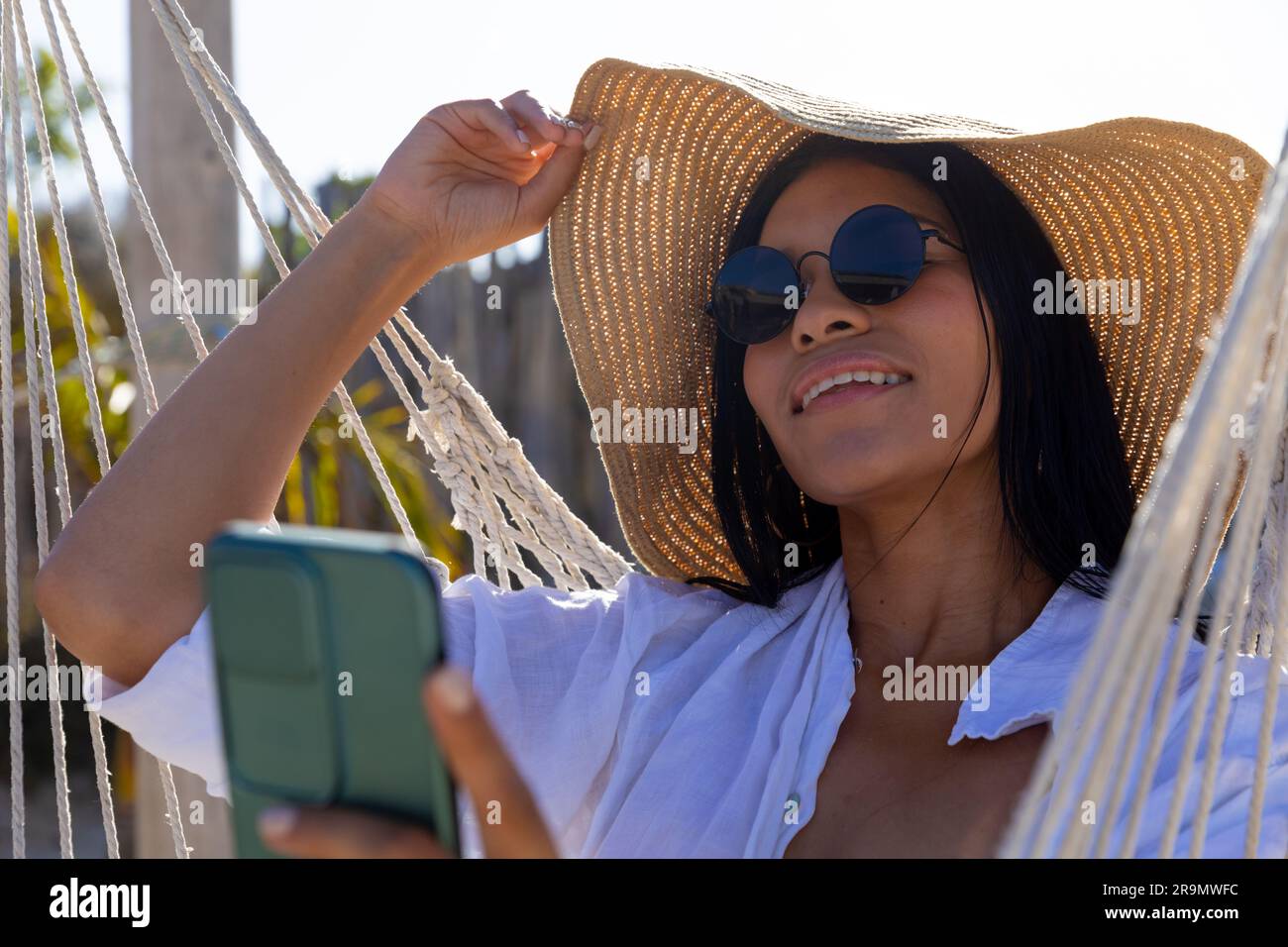 Felice donna birazziale con occhiali da sole e cappello seduto su un'amaca che scatta selfie sulla spiaggia soleggiata Foto Stock