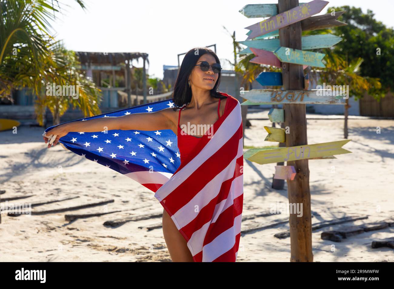Felice donna birazziale con occhiali da sole e costume da bagno rosso che balla con bandiera americana sulla spiaggia soleggiata Foto Stock