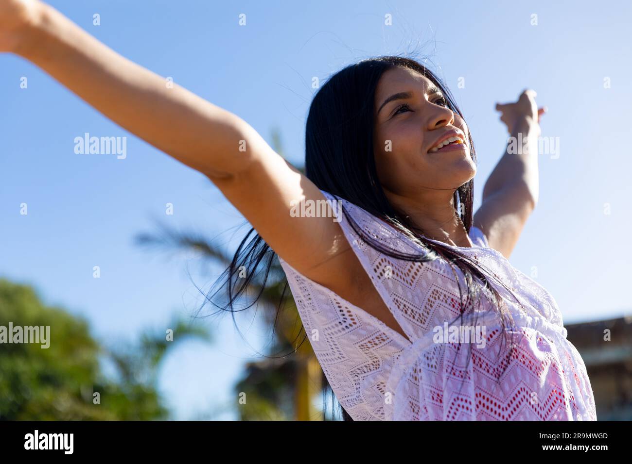 Felice donna birazziale in abito da spiaggia bianco che balla sulla spiaggia soleggiata sorridendo Foto Stock