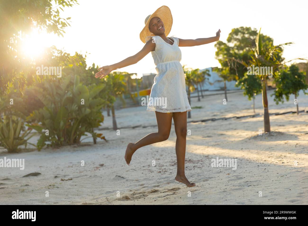 Ritratto di una donna afroamericana felice con cappello e abito da spiaggia bianco che balla sulla spiaggia soleggiata Foto Stock
