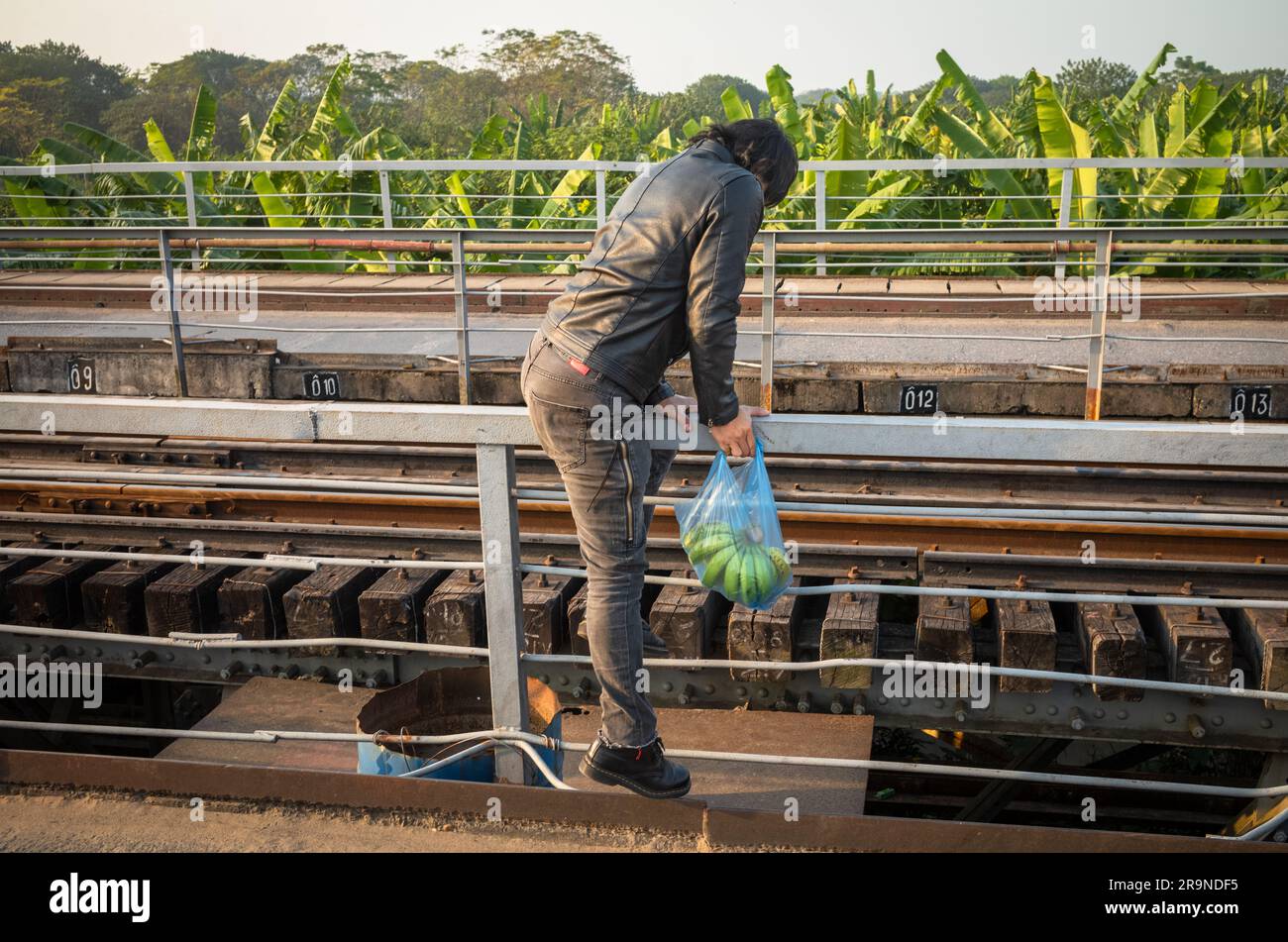 Un uomo vietnamita che indossa una giacca di pelle e porta con sé una borsa di banane si arrampica attraverso i binari ferroviari sul Long Bien Bridge, Hanoi, Vietnam. Foto Stock