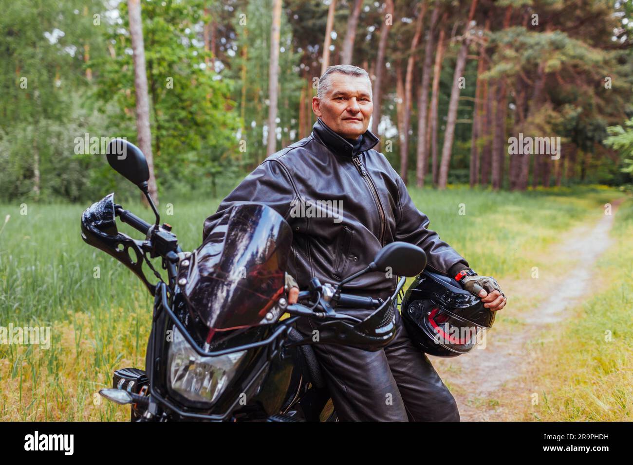 Biker anziano appoggiato su motobike nella foresta estiva. Uomo in pelle vestiti in piedi con le braccia incrociate godendo la natura. dopo il giro Foto Stock