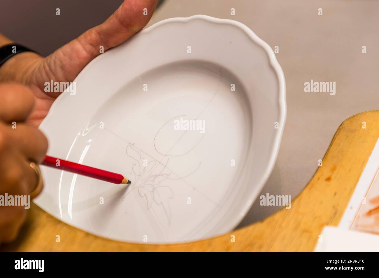 Una mano disegna il contorno di un motivo floreale con una matita rossa su un piatto di porcellana bianca non dipinto nell’officina dimostrativa del Museo della Porcellana della Fondazione Porcellana di Meissen, Sassonia, Germania. Foto Stock