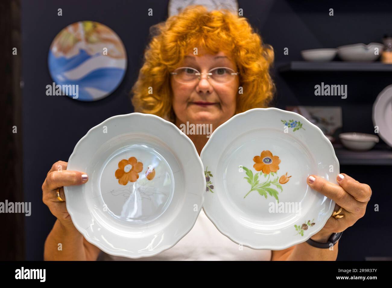 Un'artigiana tiene in mano due piatti di porcellana finiti decorati con un motivo a fiori arancioni – uno completamente dipinto, l'altro che mostra ancora il disegno a matita. La fotografia è stata scattata nell'officina dimostrativa del Museo della Porcellana della Fondazione della Porcellana di Meissen, Sassonia, Germania. Foto Stock