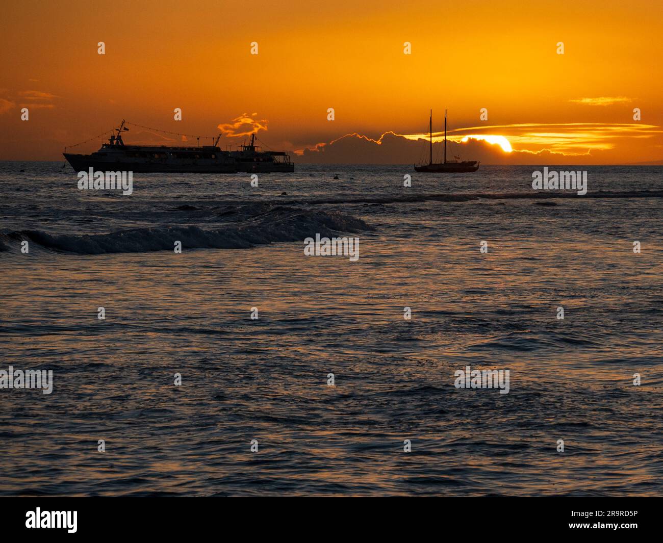 Il tramonto sull'Oceano Pacifico visto da una spiaggia di Maui brilla di arancione e mette in evidenza le onde e le navi di passaggio Foto Stock