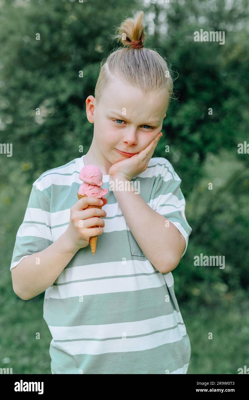 Un bambino triste stringeva la guancia con la mano e teneva un cono gelato nell'altra mano. Problemi di sensibilità dei denti in un bambino o di decadimento dei denti. Foto Stock