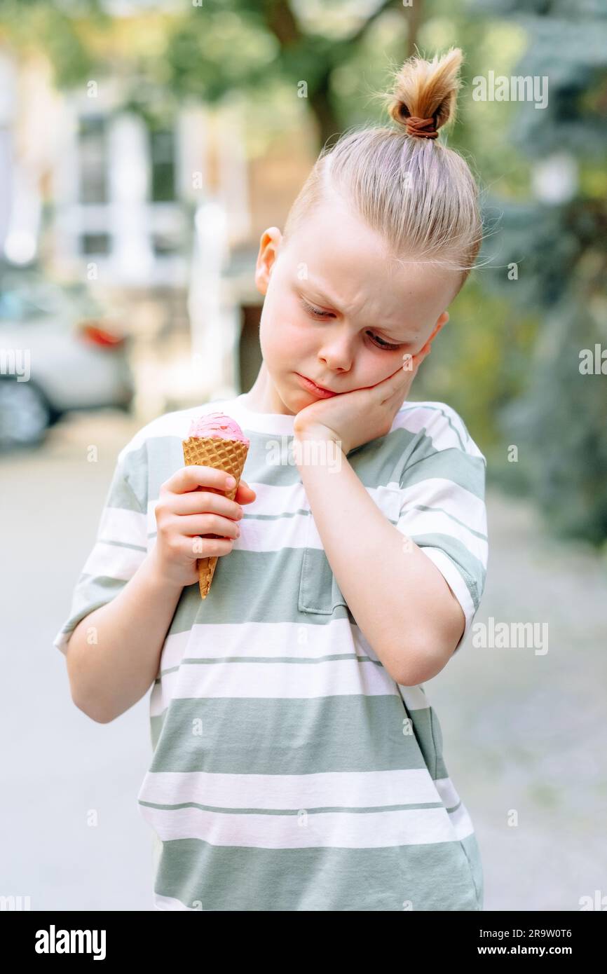 Un bambino triste stringeva la guancia con la mano e teneva un cono gelato nell'altra mano. Problemi di sensibilità dei denti in un bambino o di decadimento dei denti. Foto Stock