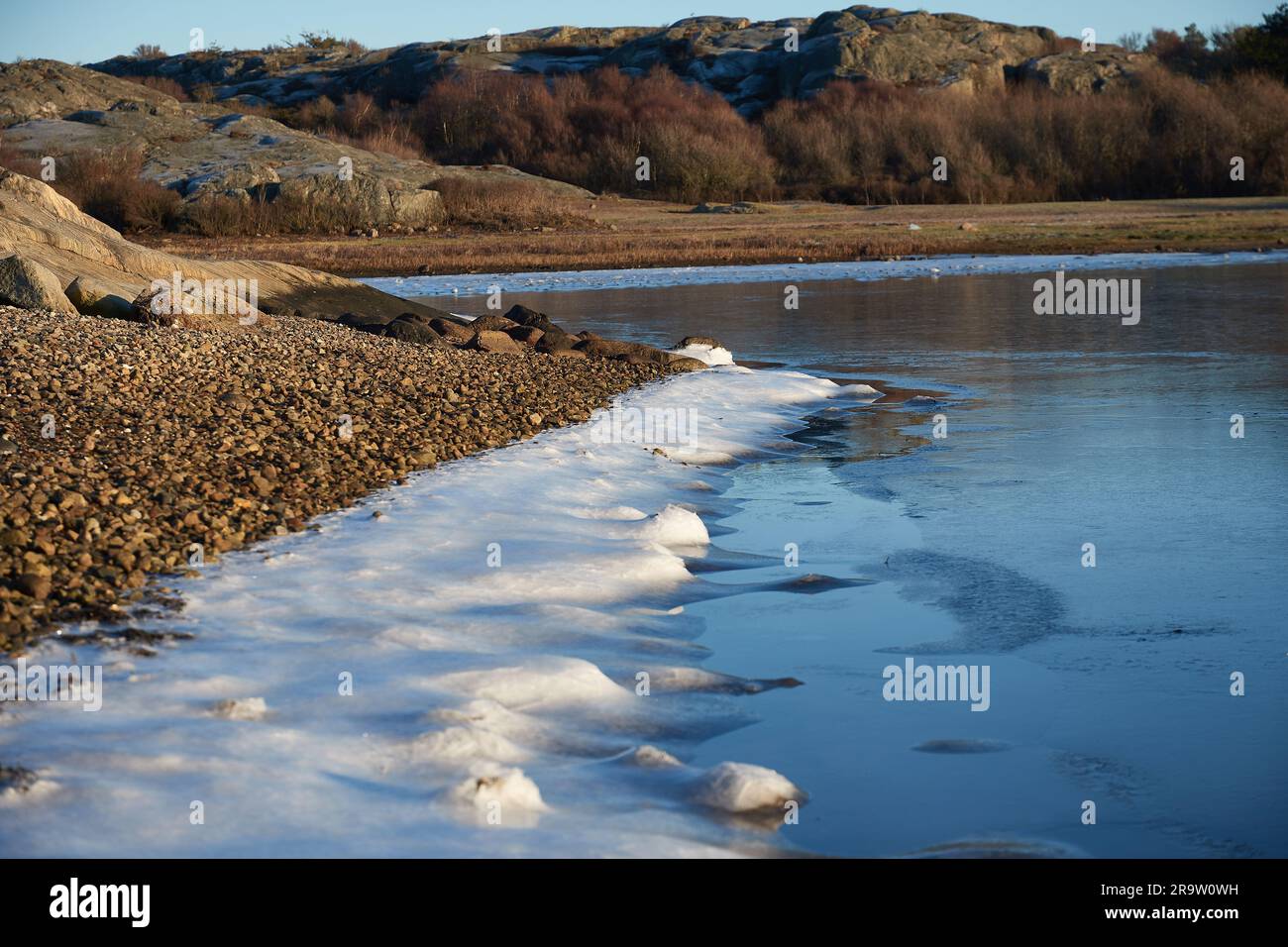 Un lago ghiacciato con ghiaccio e ghiaia sulla riva in primo piano e sauri sullo sfondo Foto Stock