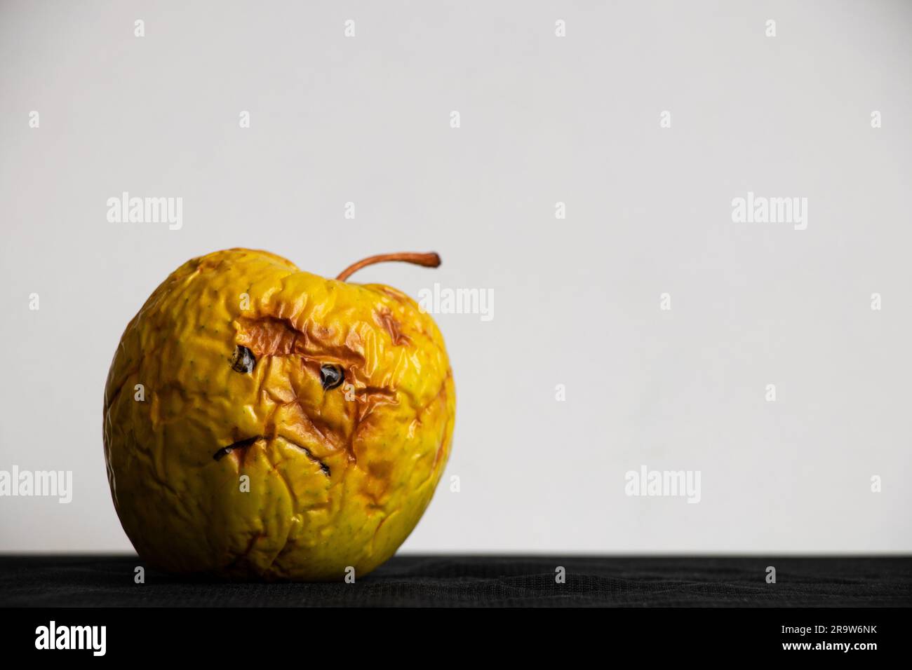 Una mela marcio e lenta con un triste sorriso dipinto su uno sfondo bianco su un tavolo nero, di cattivo umore Foto Stock