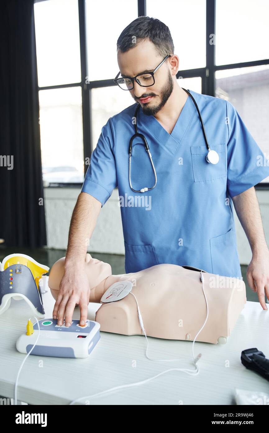 Operatore sanitario in uniforme blu, stetoscopio e occhiali che utilizza il defibrillatore automatico vicino al manichino RCP, apprendimento pratico di pronto soccorso e crit Foto Stock