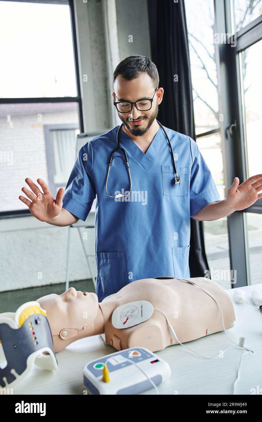 Paramedico con barba sorridente in uniforme blu e occhiali che gestiscono vicino al manichino RCP con defibrillatore nella sala di addestramento, pronto soccorso apprendimento pratico di un Foto Stock