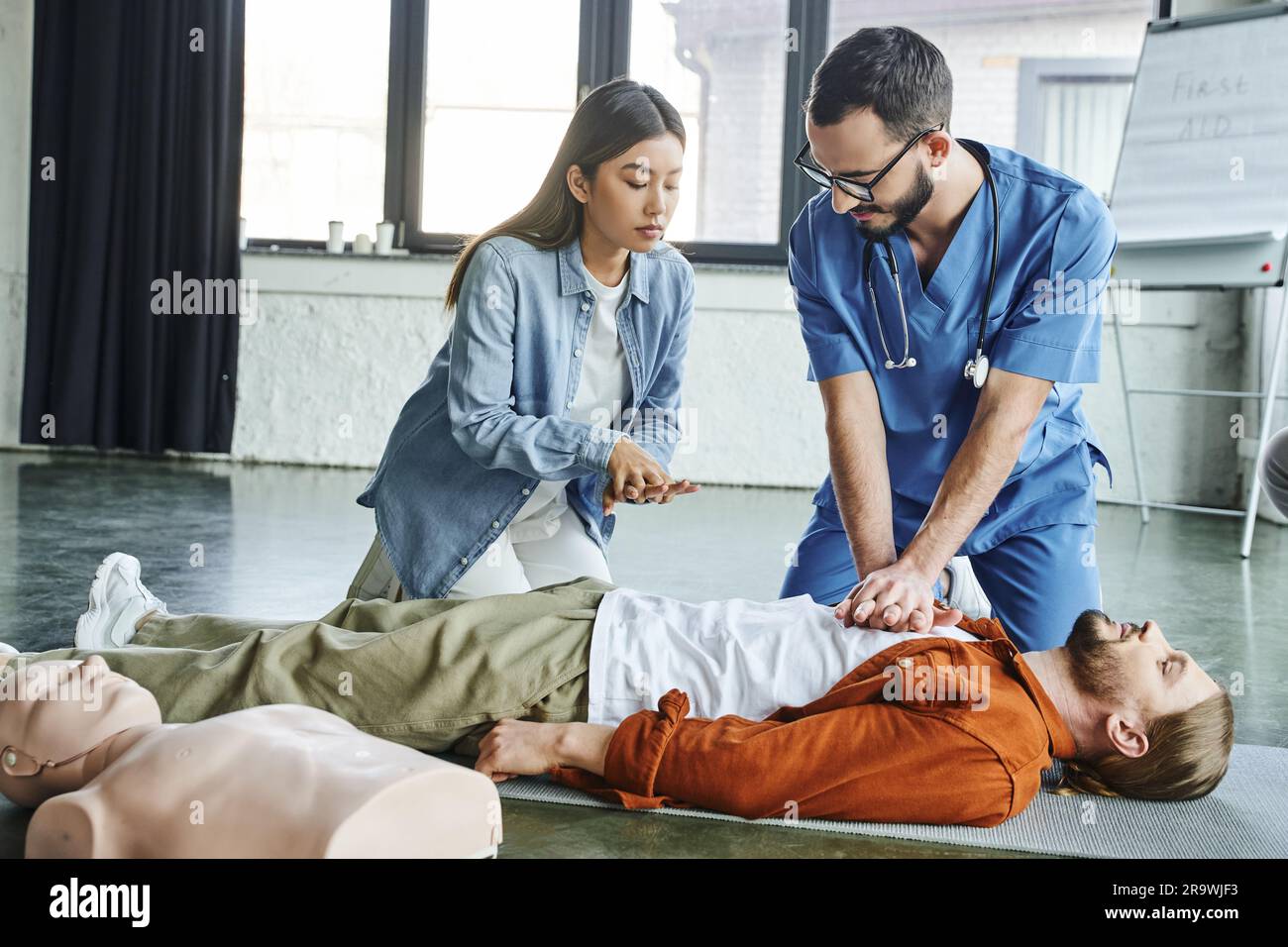 seminario medico, operatore sanitario in uniforme e occhiali da vista mostrando alle donne asiatiche tecniche di rianimazione cardiopolmonare su uomo sdraiato in formazione Foto Stock