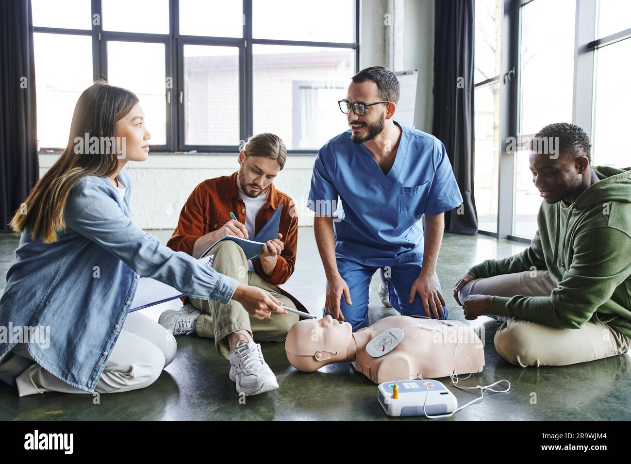 Apprendimento pratico di pronto soccorso, donna asiatica con clipboard che punta con penna al manichino RCP con defibrillatore vicino a uomini paramedici e multiculturali, guarisce Foto Stock