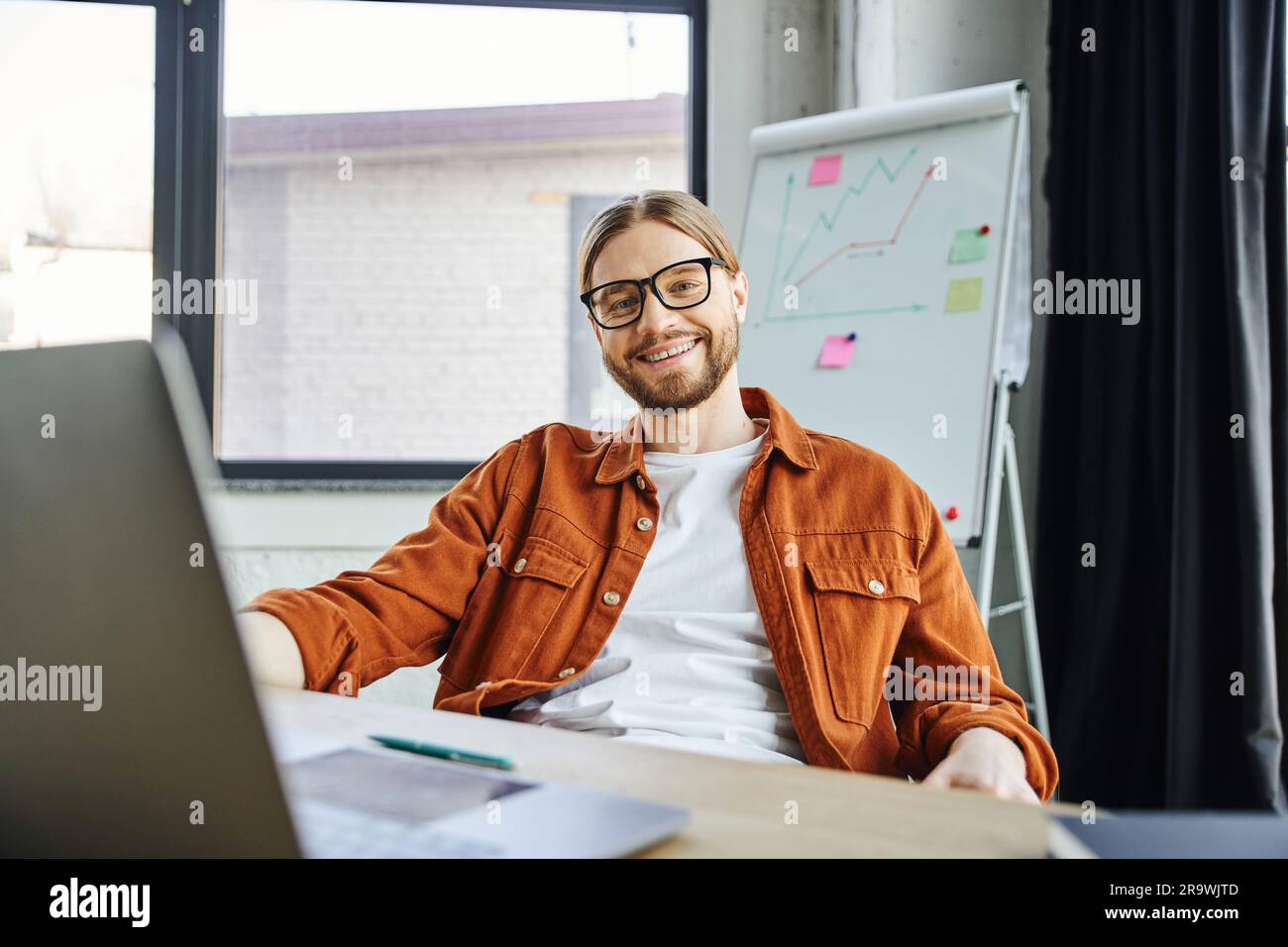 uomo d'affari barbuto felice e ottimista con occhiali da vista e camicia alla moda seduto al laptop e guardando la fotocamera accanto alla lavagna a fogli mobili sullo sfondo sfocato Foto Stock