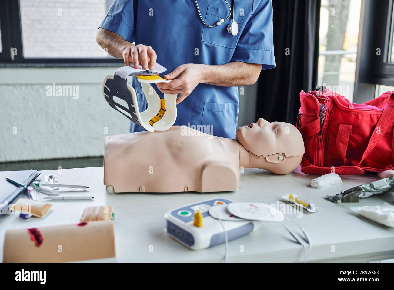 Vista parziale dell'operatore sanitario in uniforme blu che tiene il braccio del collo sopra il manichino RCP, il kit di pronto soccorso, il defibrillatore e i dispositivi medici in addestramento r Foto Stock