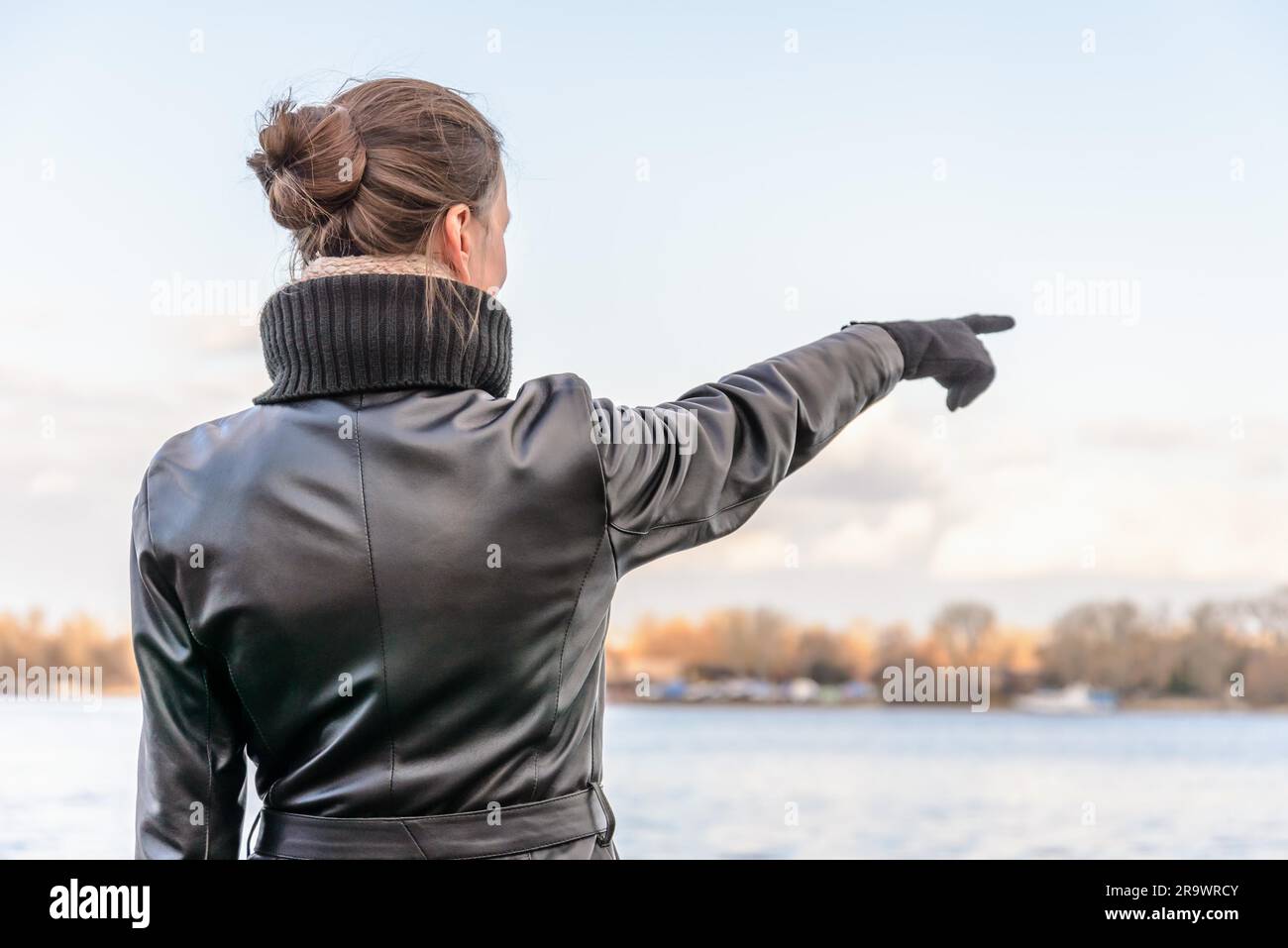 Un adulto con un chignon e indossa un cappotto di pelle nera rimane vicino al fiume e punta il dito per indicare qualcosa di interessante sul Foto Stock