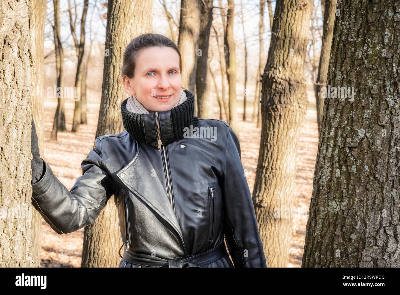 Una donna adulta che indossa una pelle nera di rivestimento è in piedi in rovere e foresta di alberi Foto Stock