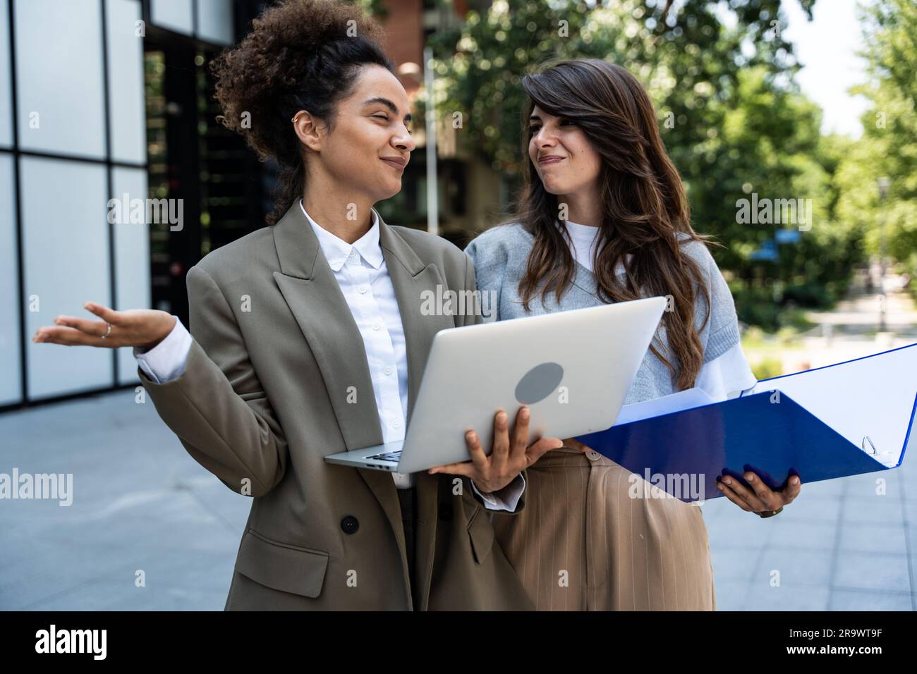 Due donne d'affari che visualizzano i dati su un notebook e statistiche nel registro. Giovani colleghi che si incontrano fuori dall'edificio degli uffici discutendo di excha Foto Stock