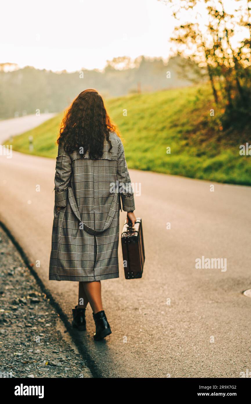 Bella donna che si allontana lungo la strada, indossa un lungo cappotto, regge una vecchia valigia vintage, vista sul retro Foto Stock