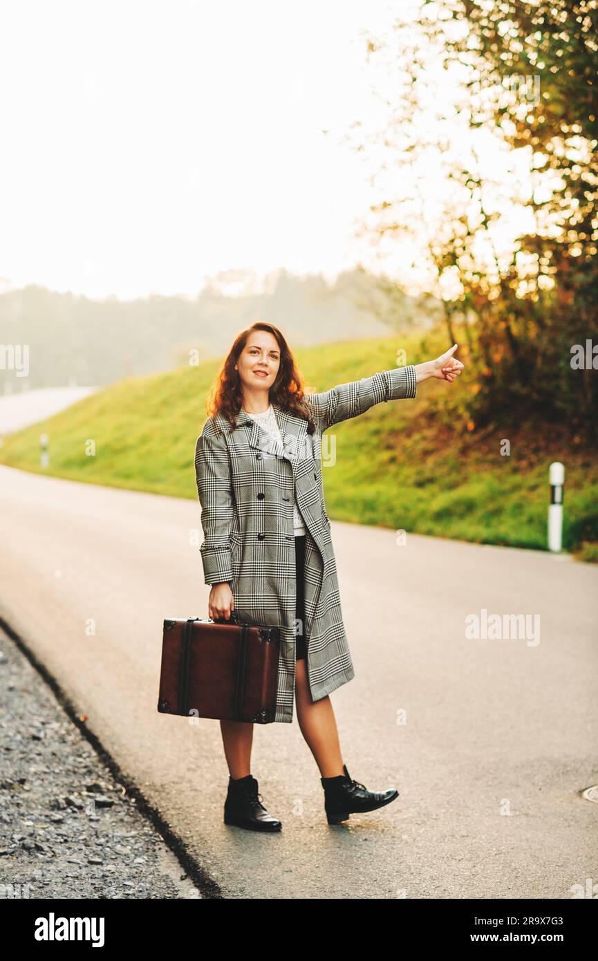 Bella donna che si allontana lungo la strada, indossa un lungo cappotto e tiene in mano una vecchia valigia vintage Foto Stock