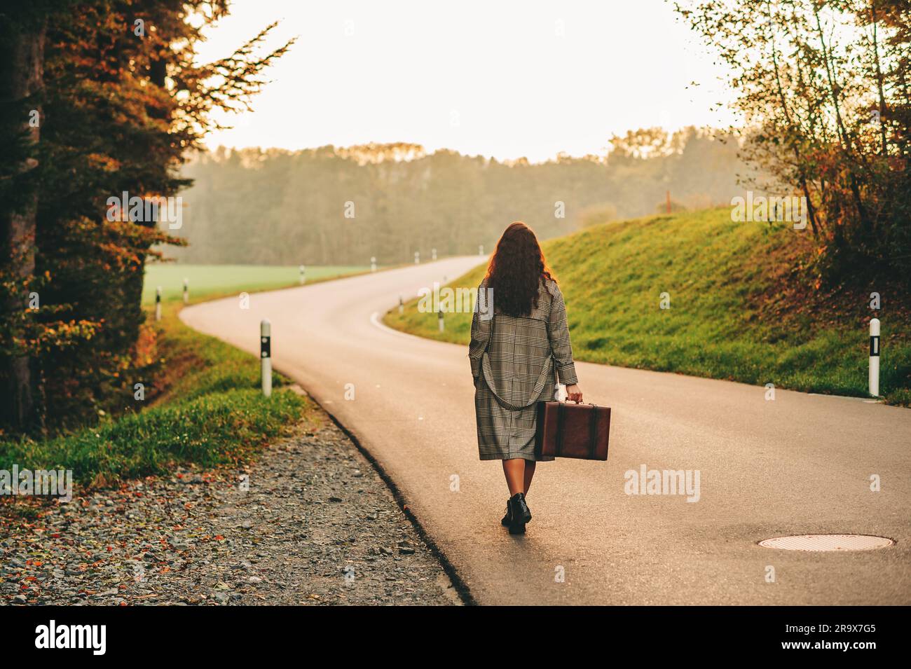 Bella donna che si allontana lungo la strada, indossa un lungo cappotto, regge una vecchia valigia vintage, vista sul retro Foto Stock