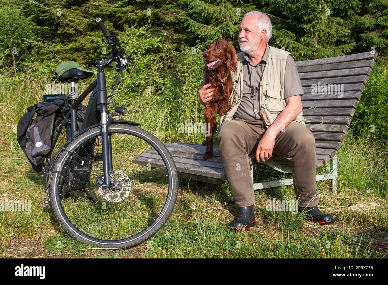 La mattina presto, un uomo anziano sta pedalando attraverso la foresta e sta facendo una pausa su una panchina di legno. Foto Stock