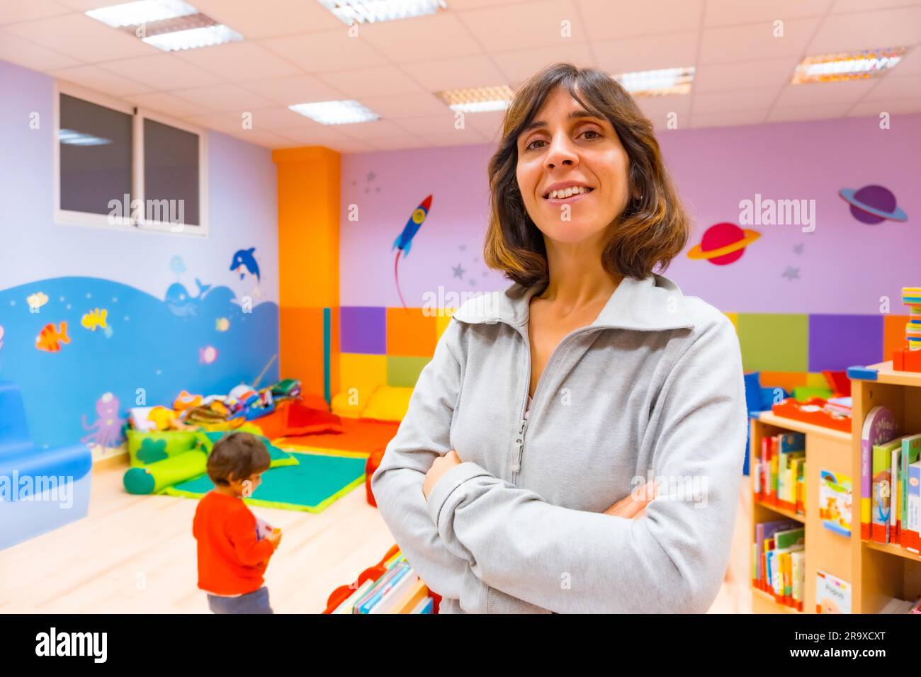 Ritratto di un asilo o di un insegnante di educazione della prima infanzia all'interno di un asilo, con un bambino che gioca sullo sfondo Foto Stock