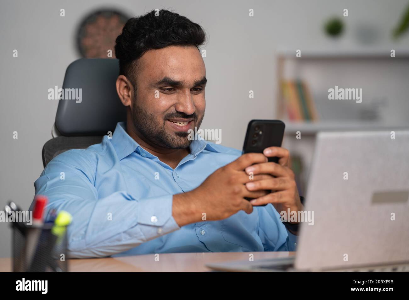 Felice e sorridente dipendente indiano che utilizza il telefono cellulare davanti al laptop in ufficio - concetto di pausa, relax e tecnologia. Foto Stock
