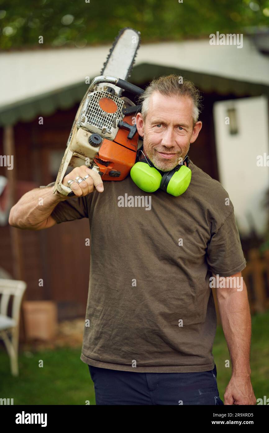 Giardiniere di mezza età che tiene una motosega sulla spalla dopo il lavoro con protezione per le orecchie che guarda la fotocamera Foto Stock