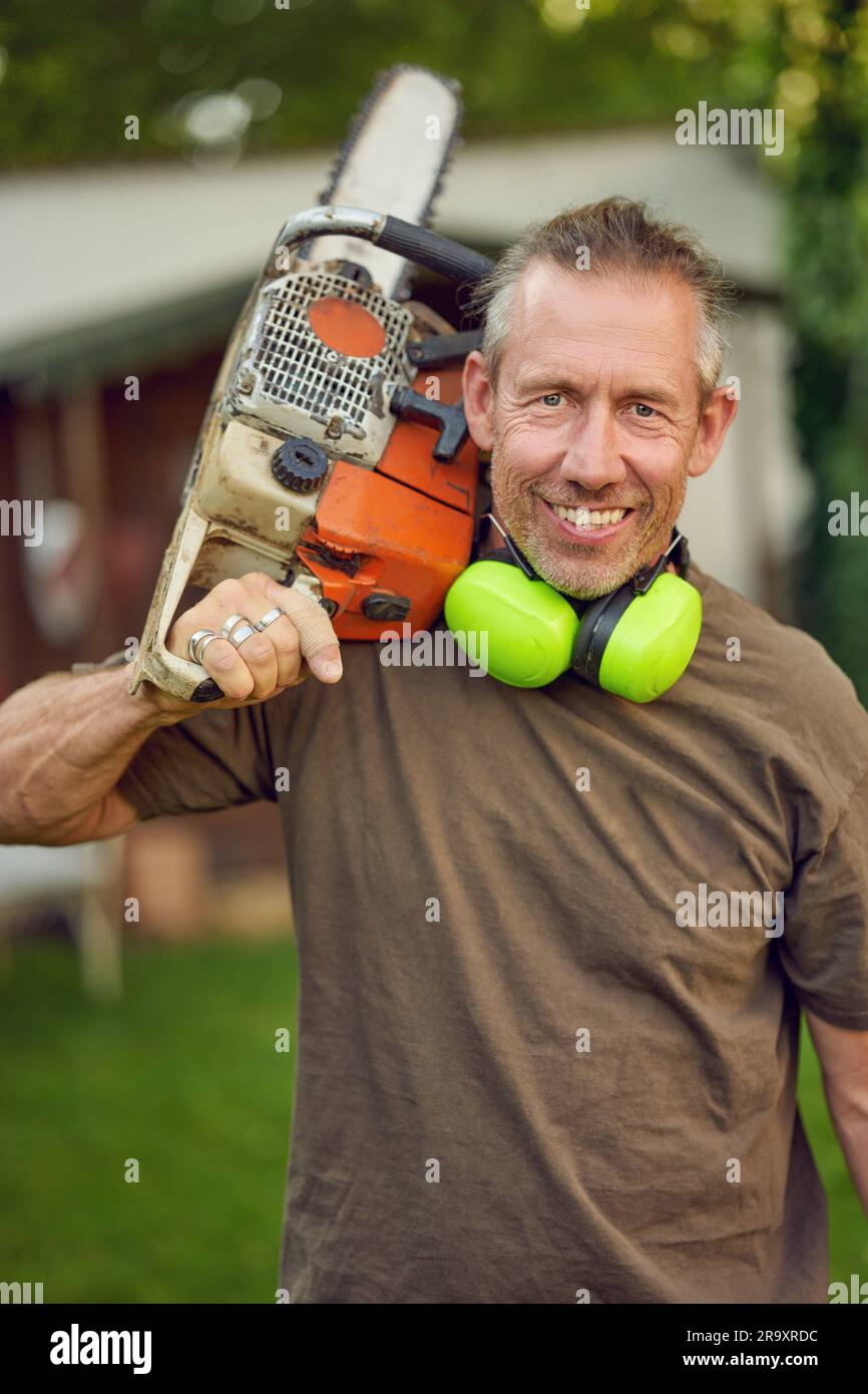 Giardiniere di mezza età che tiene una motosega sulla spalla dopo il lavoro con protezione per le orecchie che guarda la fotocamera Foto Stock