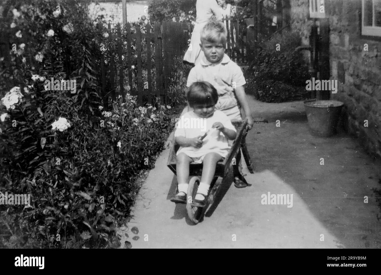 Un ragazzo e una ragazza in un giardino, seduto in una ruota di legno barrow mentre il ragazzo la spinge, Surrey, c1930. Si tratta di una fotografia tratta da un album di scatti, durante l'occupazione della Palestina da parte dell'esercito britannico, con foto provenienti dalla Palestina e dal Regno Unito, tra cui devil's Jump vicino a Churt nel Surrey e devil's Punch Bowl. L'addestramento sembra essere a Hursley Camp, vicino a Winchester. La Gran Bretagna amministrò la Palestina per conto della società delle Nazioni tra il 1920 e il 1948, un periodo denominato "mandato britannico”. Foto Stock