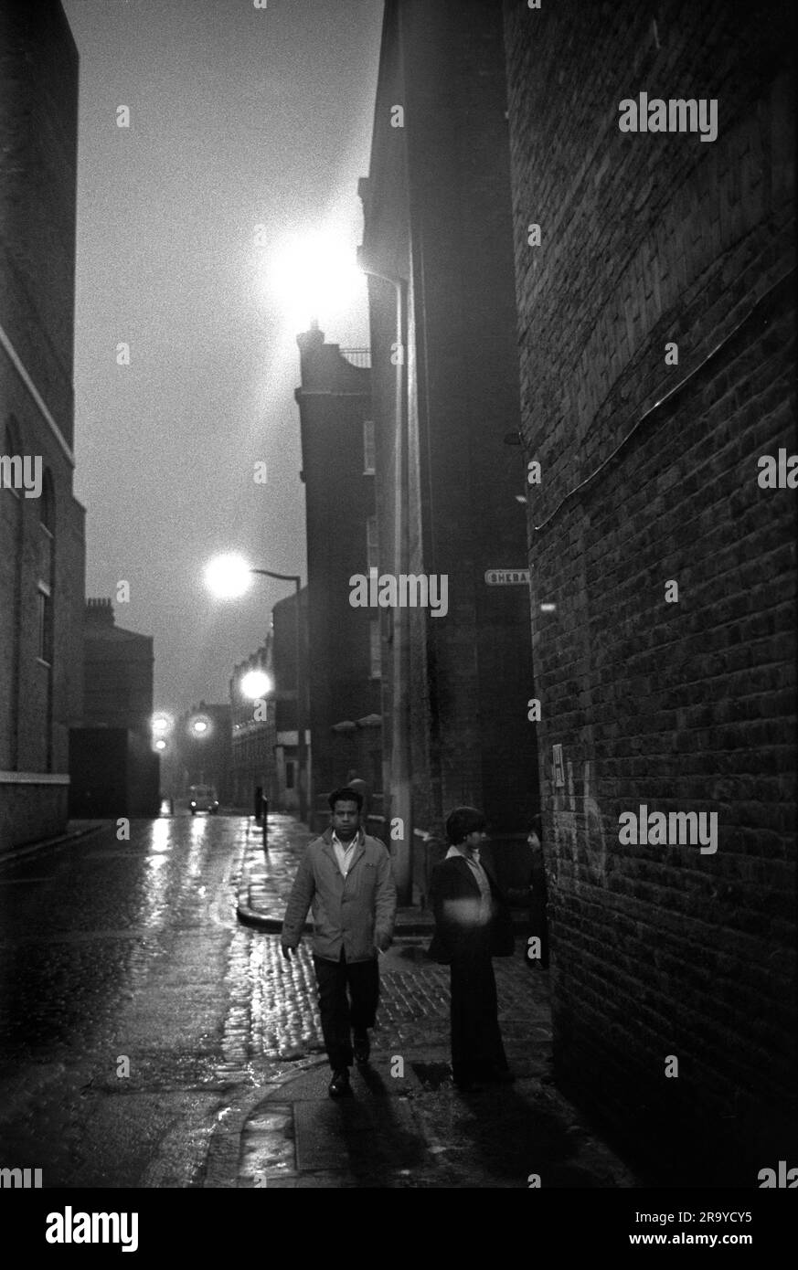 Victorian Slum Buildings East End Londra 1970s, Brick Lane all'incrocio con Sheba Street. Un uomo asiatico e un ragazzo adolescente camminano davanti ai blocchi di appartamenti vittoriani. Tower Hamlets, Londra, Inghilterra circa 1976. 70S UK HOMER SYKES Foto Stock