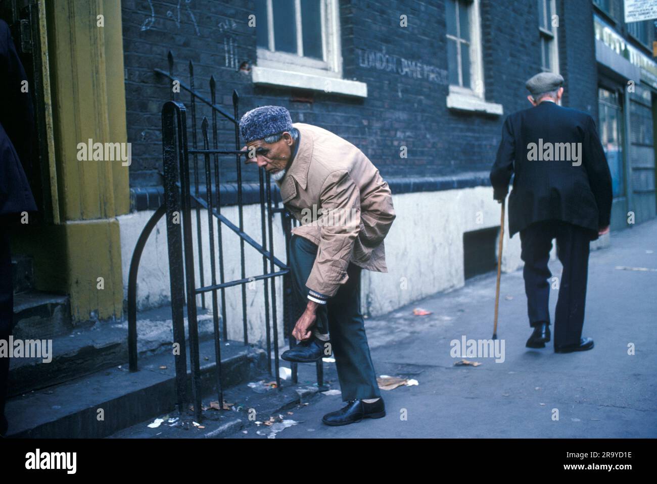 Whitechapel, Brick Lane, Londra anni '1970 Regno Unito. Un vecchio musulmano si toglie le scarpe prima di entrare nella Moschea Jamme Masjid per le preghiere del venerdì. (Formalmente la Sinagoga Machzike Hadath all'angolo tra Brick Lane e Fournier Street), mentre un anziano Cockney con bastone da passeggio e cappello piatto passa oltre Brick Lane. Tower Hamlets, East End Londra, Inghilterra 1978. HOMER SYKES anni '70 Foto Stock