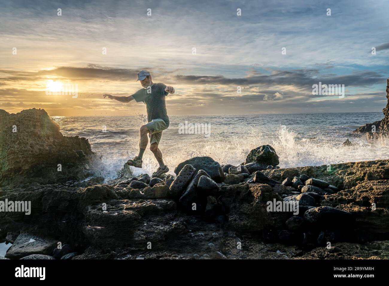 Uomo che salta nel surf su una costa piena di rocce basaltiche da un antico flusso lavico mentre il sole sorge dietro le nuvole, Poipu, Kauai, Hawaii Foto Stock