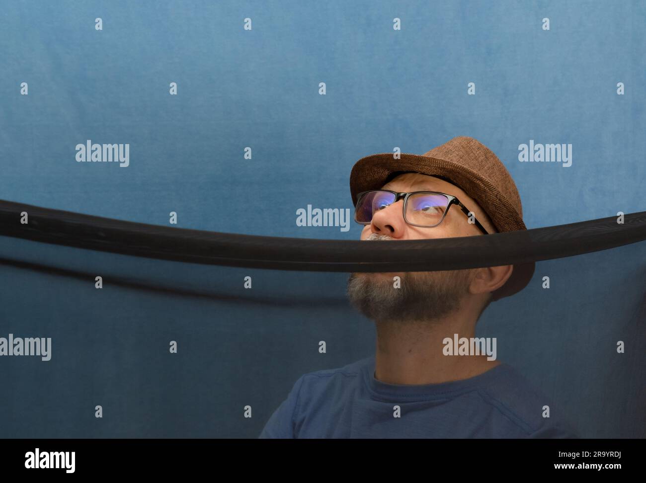 Uomo che indossa un cappello, occhiali e barba dietro un panno trasparente di colore nero. isolato su sfondo blu. Foto Stock