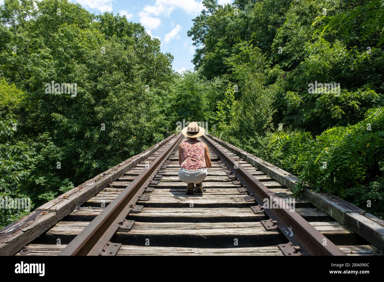 Vista posteriore di una donna caucasica sui binari ferroviari nei boschi di campagna in una giornata di sole. Foto Stock