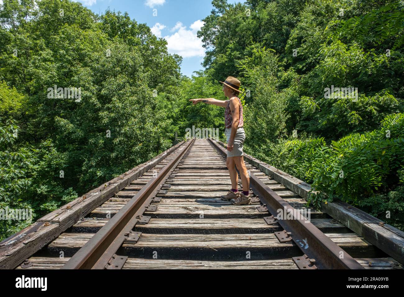 Vista laterale di una donna caucasica sui binari ferroviari nei boschi di campagna in una giornata di sole. Foto Stock