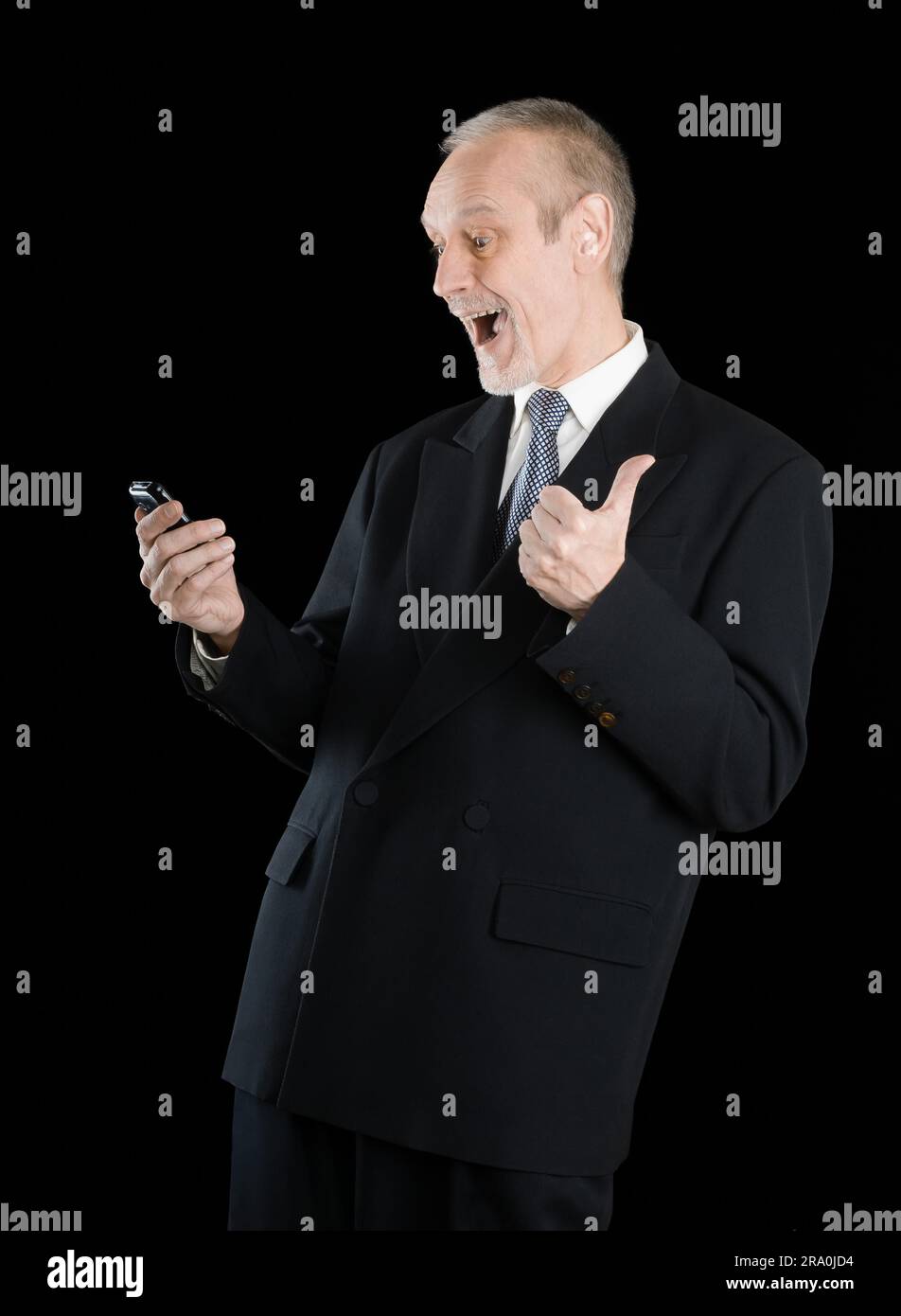 Happy businessman indossando un vestito nero, sorridente e la lettura di sms sul cellulare, con pollice in alto, su sfondo nero Foto Stock
